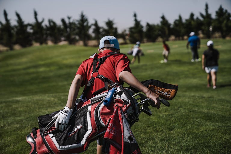 A junior golfer in red carrying a bag on a lush green golf course during a sunny day.
