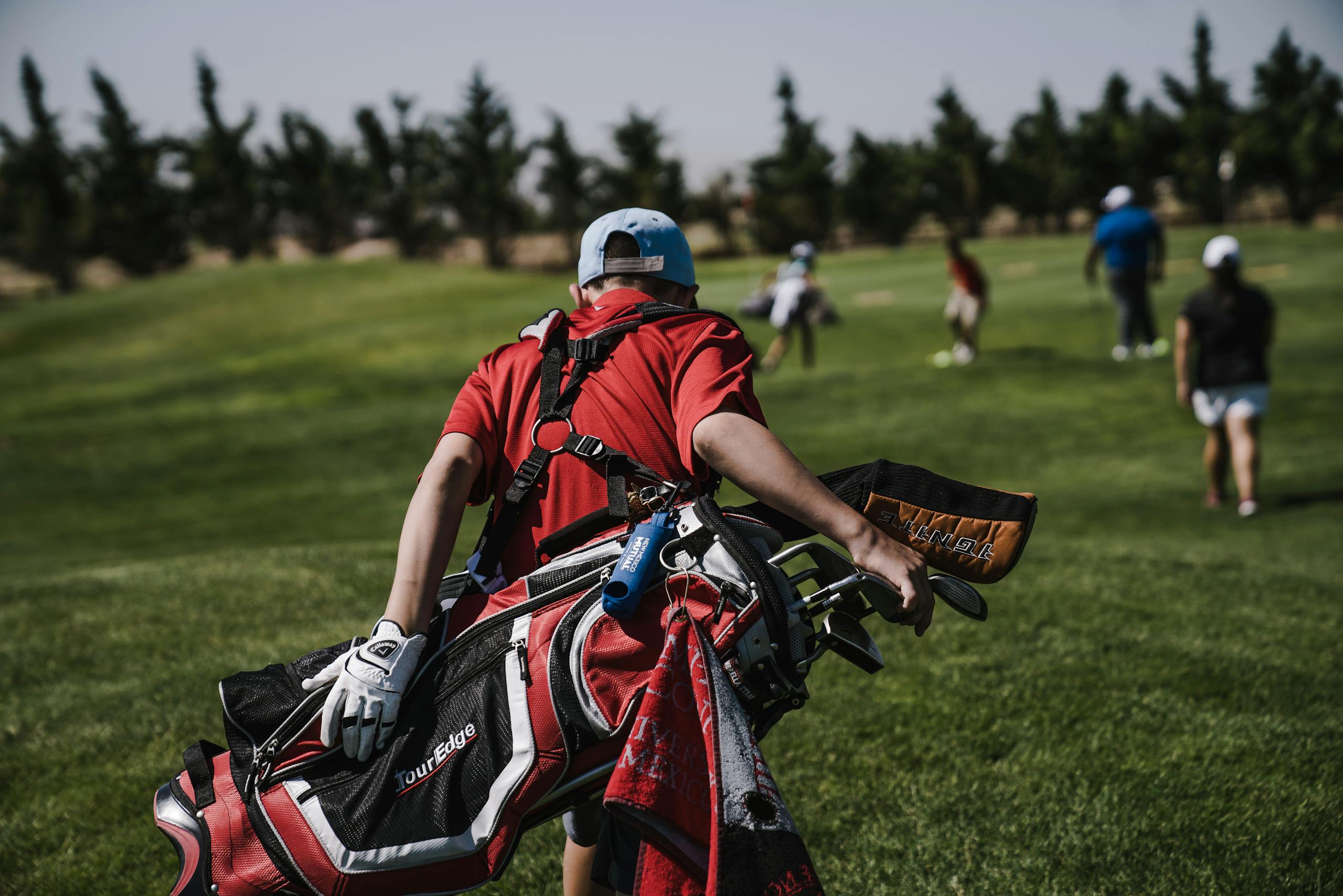 A junior golfer in red carrying a bag on a lush green golf course during a sunny day.